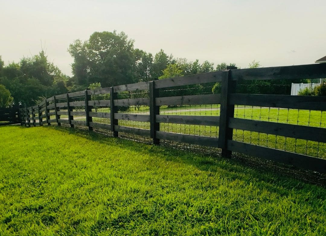 A black wooden fence runs along a lush green field under a clear sky.